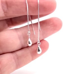 May include: A pair of silver threader earrings featuring a delicate box chain design that suspends a small, smooth, polished teardrop-shaped charm at the end of each chain, creating a simple and reflective metallic appearance.