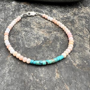 May include: A delicate bracelet featuring a strand of small, faceted, pale peach-colored beads accented with a short segment of cylindrical, bright turquoise-colored beads. The piece is finished with a silver-toned metal lobster claw clasp.