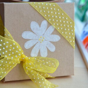 K&ouml;nnte beinhalten: Eine braune Geschenkbox mit einem gelben gepunkteten Band und einer wei&szlig;en G&auml;nsebl&uuml;mchen-Malerei auf der Vorderseite. Die Box ist mit einem gelben gepunkteten Band umwickelt.