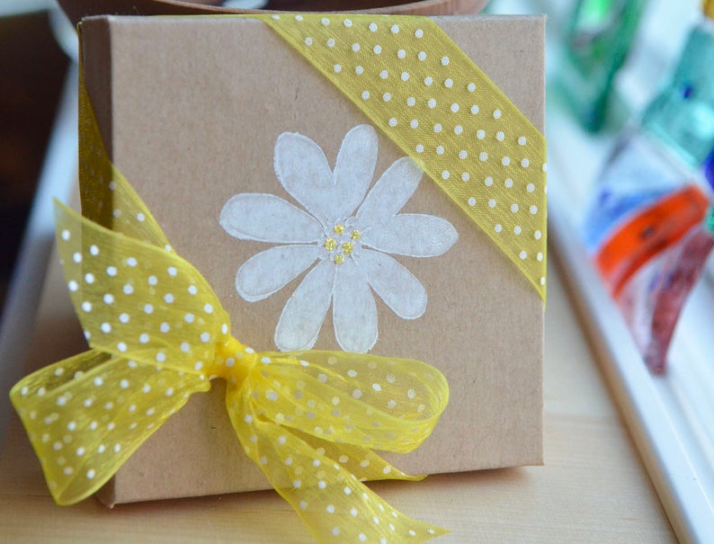 K&ouml;nnte beinhalten: Eine braune Geschenkbox mit einem gelben gepunkteten Band und einer wei&szlig;en G&auml;nsebl&uuml;mchen-Malerei auf der Vorderseite. Die Box ist mit einem gelben gepunkteten Band umwickelt.