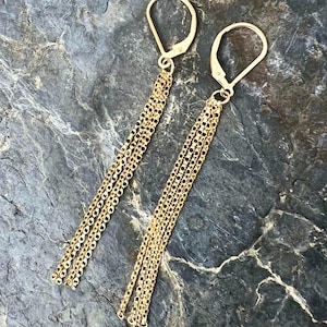 May include: Gold-toned dangle earrings. Each earring features a lever-back closure and multiple strands of delicate chain. The chains cascade downwards, creating a flowing, elegant design. The earrings are displayed against a textured, dark gray surface.