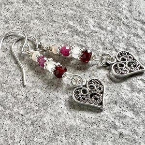 Może przedstawiać: Srebrne kolczyki wiszące z ażurowym sercem. Kolczyki posiadają sznur małych, kolorowych koralików w odcieniach różu, bieli i czerwieni. Kolczyki są prezentowane na teksturowanej szarej powierzchni.