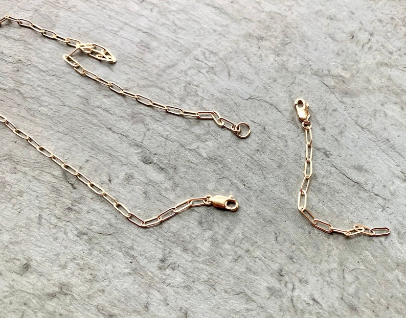 May include: Three gold chain links, each with a clasp, are laid out on a gray textured surface. The chains are made of small, oval-shaped links.