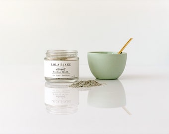 Könnte beinhalten: Ein klares Glas mit der Aufschrift "LOLA JANE antioxidant FACIAL MASK" mit dem Text "SEA CLAY + SPIRULINA". Ein kleiner Haufen graugrünes Pulver, eine hellgrüne Schüssel und ein goldfarbener Löffel sind ebenfalls vorhanden. Die Artikel sind auf einer reflektierenden weißen Oberfläche angeordnet.