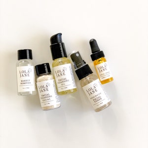 May include: A collection of Lola Jane skincare products, including makeup remover, facial cleansing grains, facial cleansing oil, rosewater facial toner, and facial serum. Each product is in a small, clear bottle with a black cap or pump. The bottles are arranged on a white surface.