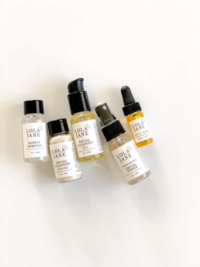 May include: A collection of Lola Jane skincare products, including makeup remover, facial cleansing grains, facial cleansing oil, rosewater facial toner, and facial serum. Each product is in a small, clear bottle with a black cap or pump. The bottles are arranged on a white surface.