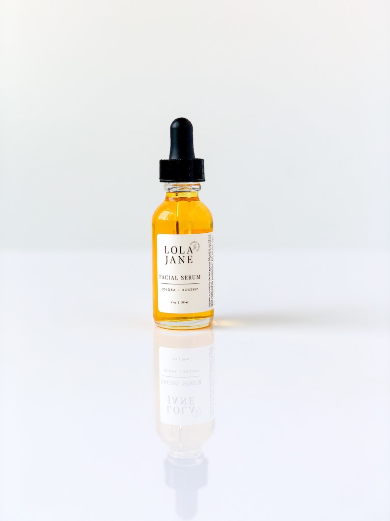 Puede incluir: Una botella de vidrio transparente de suero facial Lola Jane con un cuentagotas negro. La botella contiene un l&iacute;quido de color dorado y tiene una etiqueta blanca con texto negro. La etiqueta dice "Facial Serum" y "Jojoba + Rosehip".