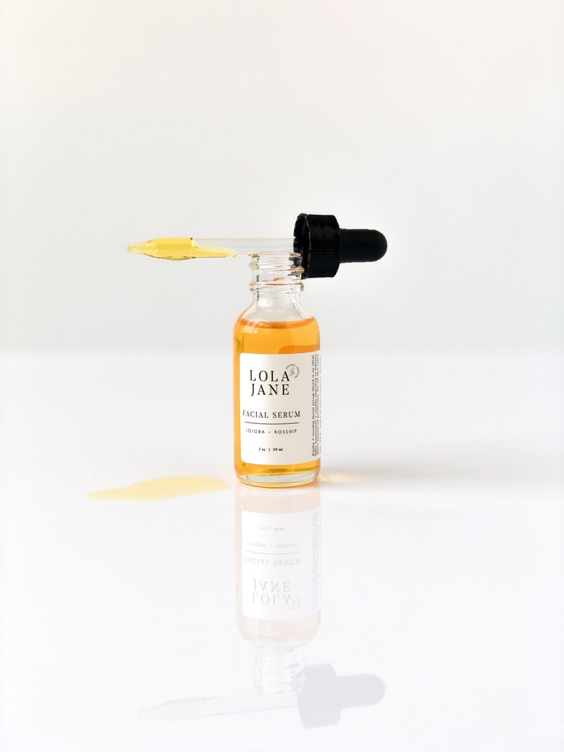 Puede incluir: Una botella de vidrio transparente de suero facial Lola Jane con un cuentagotas negro. La botella contiene un l&iacute;quido amarillo y tiene una etiqueta blanca con texto negro. El cuentagotas est&aacute; lleno de suero, y se ha derramado un poco.