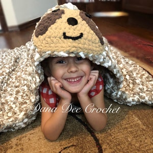 May include: A child wearing a red and white checkered shirt is lying on a brown and white crocheted blanket with a sloth face hood. The blanket is made by Dana Dee Crochet.