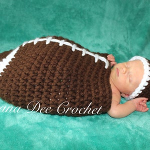 May include: A brown and white crocheted football-shaped baby blanket and matching hat. The blanket has white stitching details. The text "Dana Dee Crochet" is visible in the bottom left corner of the image.