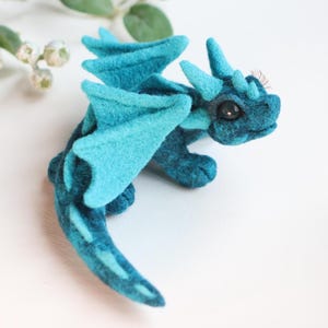 Può includere: Statuetta di drago in feltro fatta a mano, nei toni del verde acqua e blu. Il drago ha ali, corna e una lunga coda dettagliate. La statuetta è posizionata su una superficie bianca, con un rametto di verde sullo sfondo.
