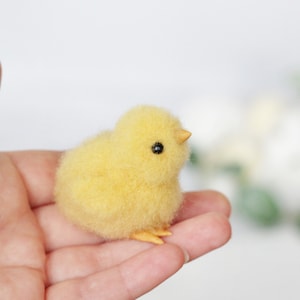 May include: A small, yellow, fluffy chick with black eyes. The chick is sitting on a person's hand.
