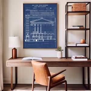 May include: A blueprint style print of the New York Stock Exchange building, framed in a dark wood frame and hanging on a white wall above a wooden desk with a brown leather chair. The print is on a blue background with white lines and text. The text reads "NEW YORK STOCK EXCHANGE BROAD STREET ELEVATION".