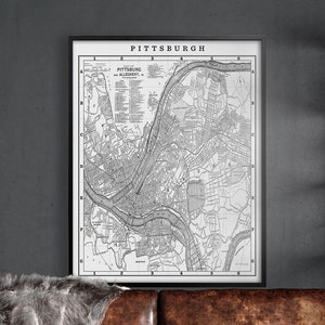 May include: A brown leather sofa with a gray faux fur throw blanket. A vintage map of Pittsburgh, Pennsylvania hangs on the wall above the sofa. Two industrial metal shelves are on either side of the sofa.