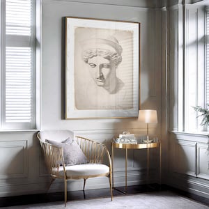 May include: A framed print of a classical bust in a gold frame hangs on a wall. The artwork is a sepia-toned sketch of a woman's face. A gold side table and chair are in front of the wall.