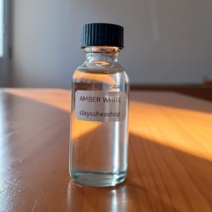 May include: A clear glass bottle with a black cap filled with a clear liquid. The bottle has a white label that reads "AMBER WHITE clayssheashop".