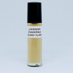 May include: A clear glass roller bottle filled with a light beige liquid, topped with a black cap. A white rectangular label on the front features the black text: LAVENDER, CHAMOMILE, YLANG-YLANG. The bottle stands upright against a plain background.