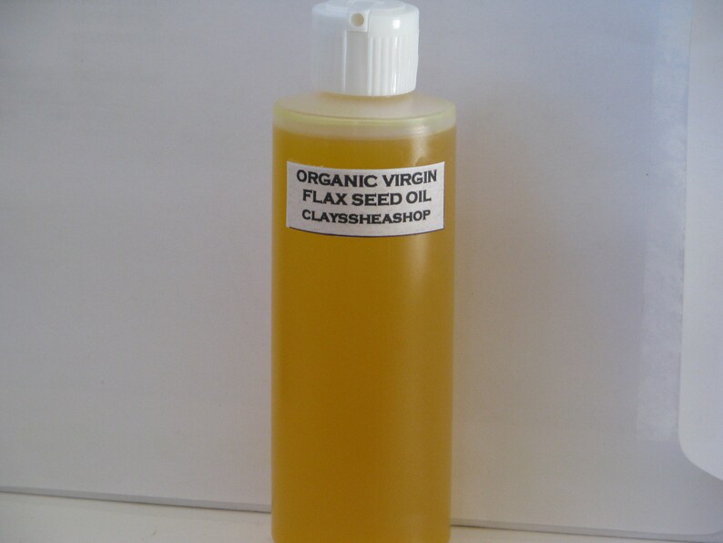 May include: A clear glass bottle filled with golden yellow organic virgin flax seed oil. The bottle has a white plastic cap and a white label with black text that reads "ORGANIC VIRGIN FLAX SEED OIL CLAYSSHEASHOP".