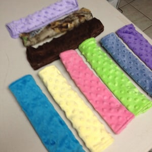 May include: Assortment of colorful fabric car seat belt covers. The covers are made of soft, plush fabric with a textured dot pattern. The colors include light blue, yellow, pink, green, purple, and brown.
