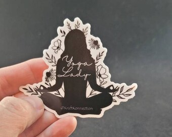 May include: Black and white silhouette of a person in a yoga pose with floral accents. The text "Yoga Lady" is written in cursive above the figure. The text "@KraftKonnection" is written below the figure.