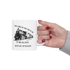 Op de afbeelding: Witte keramische mok met een zwart-witte illustratie van een stoomtrein en de tekst "Dwing me niet om je naar het station te brengen".