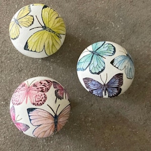 May include: Three round cabinet knobs with a white base. The top left knob features yellow butterflies. The right knob displays light blue and purple butterflies. The bottom left knob is decorated with pink and peach-coloured butterflies.