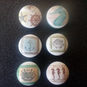 Può includere: Set di sei pomelli rotondi in ceramica con temi nautici. I pomelli presentano immagini di una stella marina, una ruota, un cavalluccio marino, un'ancora, un pesce e tre cavallucci marini.