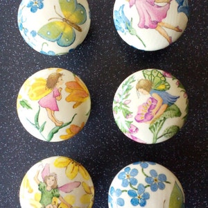 May include: Six round ceramic knobs with a white background and colourful floral and fairy designs. The knobs feature butterflies, flowers, and fairies in various colours, including pink, blue, yellow, and green.