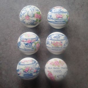 Può includere: Sei pomelli in ceramica bianca con motivi floreali blu e rosa. I pomelli presentano tazze da tè, piattini e motivi floreali. Alcuni pomelli hanno testo in francese, tra cui "Salon du The" e "Rue Saint Charles Paris".