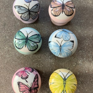 May include: Six round wooden knobs with butterfly designs. The knobs are painted in various pastel colours, including pink, blue, yellow, and green. Each knob features a different butterfly design with black outlines.