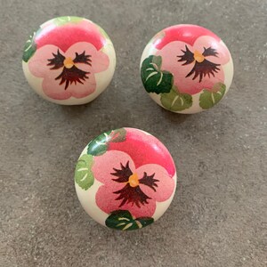 May include: Three round wooden knobs with a white background and a pink and green floral design. The flowers are pink pansies with green leaves.