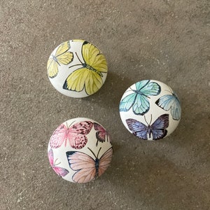 Peut inclure: Trois boutons ronds en céramique blanche avec des motifs de papillons. Les boutons présentent des papillons de différentes couleurs, notamment jaune, bleu et rose.