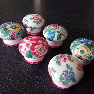 Può includere: Sei pomelli per cassetti in ceramica bianca con motivi floreali. I pomelli presentano una varietà di fiori colorati, tra cui rosso, rosa, giallo, blu e verde. Ogni pomello ha un bordo a pois rosso e bianco.