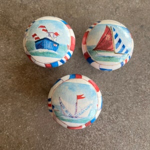 May include: Three round wooden knobs with nautical themed designs. The first knob features a blue boat with red and white flags. The second knob features a red and white striped sailboat. The third knob features a white sailboat with a red flag and the text "a life on the ocean wave".