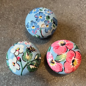 May include: Three round knobs with floral patterns. The knobs are painted a light blue colour. One knob has a pink floral pattern, one has a white floral pattern with a bee, and one has a blue floral pattern.