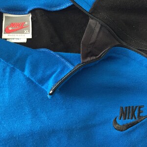 May include: A blue and black Nike quarter-zip sweatshirt with a black zipper and a black Nike logo on the front. The tag says "Nike" and "XL" and "Made in U.S.A."