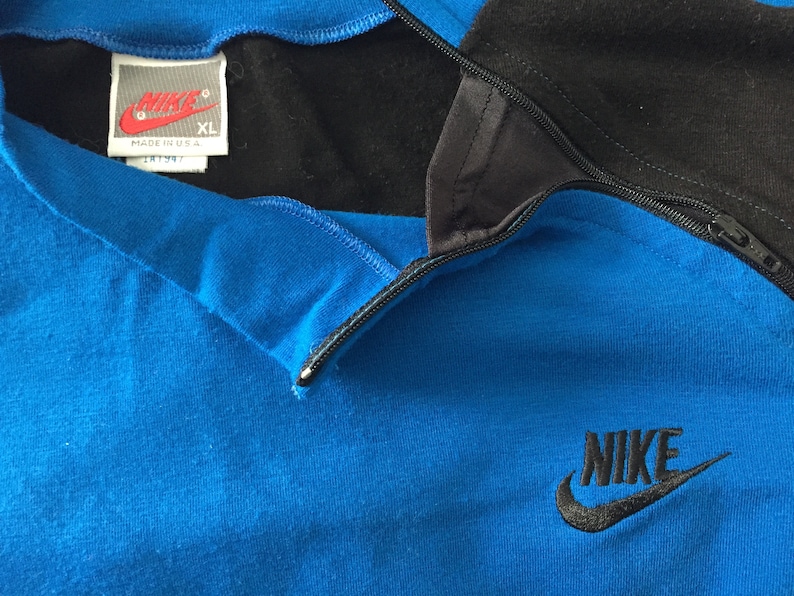 May include: A blue and black Nike quarter-zip sweatshirt with a black zipper and a black Nike logo on the front. The tag says "Nike" and "XL" and "Made in U.S.A."