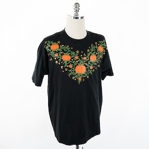 Pode incluir: Camiseta preta com um design de videira de abóbora em laranja, verde e dourado. A videira é adornada com pequenas estrelas e luas crescentes.