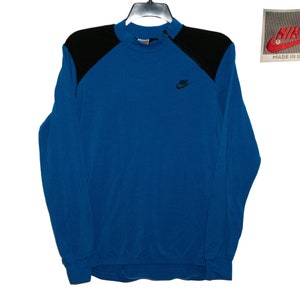 May include: A blue long-sleeve sweatshirt with black shoulder panels and a Nike logo on the front. The sweatshirt has a mock neck and a zipper on the collar. The tag says "Nike" and "Made in U.S.A."