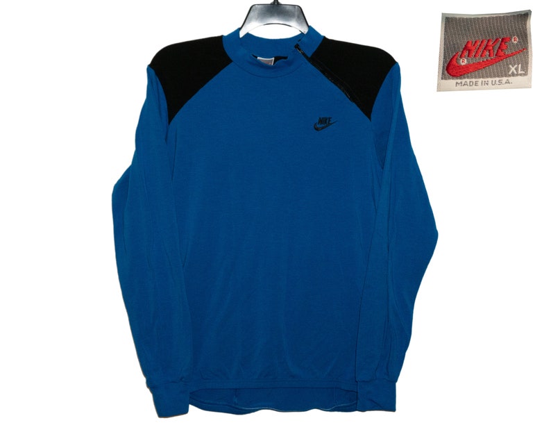 May include: A blue long-sleeve sweatshirt with black shoulder panels and a Nike logo on the front. The sweatshirt has a mock neck and a zipper on the collar. The tag says "Nike" and "Made in U.S.A."
