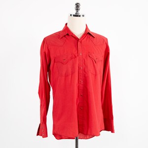 以下が含まれることがあります： 赤い長袖ボタンダウンシャツ。胸ポケットが2つと尖った襟があります。