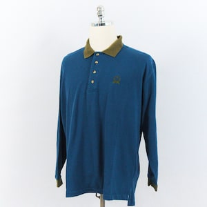 Puede incluir: Polo de manga larga azul turquesa con cuello y puños verde oliva en contraste. La camisa tiene una tapeta de tres botones y un pequeño logotipo bordado en el pecho. Se muestra en un maniquí.