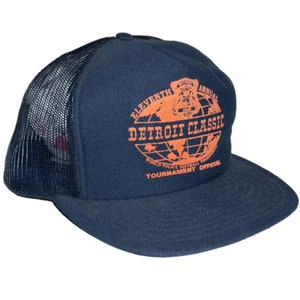 Op de afbeelding: Marineblauwe truckerpet met een mesh achterkant en een gebogen klep. De voorkant heeft een oranje afbeelding met de tekst "ELEVENTH ANNUAL DETROIT CLASSIC WORLD POLICE SOFTBALL TOURNAMENT OFFICIAL".