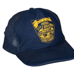 Op de afbeelding: Een donkerblauwe mesh trucker cap met een geel geborduurd patch waarop staat "Sixth Annual Detroit Classic Worldpolice Softball Tournament Aug. 15, 16, 2013"