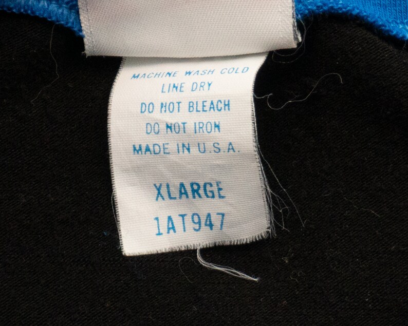 May include: White care label with black text, reading "MACHINE WASH COLD, LINE DRY, DO NOT BLEACH, DO NOT IRON, MADE IN U.S.A." Below is a white tag with black text reading "XLARGE 1AT947".