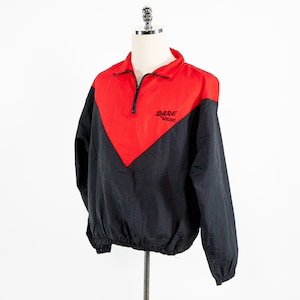 Puede incluir: Una chaqueta cortavientos negra y roja con cierre de cremallera y cordón de ajuste en la cintura. La chaqueta tiene un diseño de color contrastante con un panel rojo en la parte delantera y paneles negros en las mangas y la espalda. La chaqueta tiene la palabra "DARE" bordada en la parte delantera.