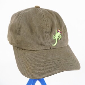 Può includere: Un cappello da baseball verde con una lucertola verde con occhiali da sole ricamata sul davanti. Il cappello è realizzato in tessuto di cotone marrone.