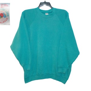 Op de afbeelding: Een turquoise blauwe trui met een ronde hals en lange mouwen. De trui heeft een Fruit of the Loom-label met de tekst "Fruit of the Loom Casualwear" en "Ladies Size 3XL 50% Cotton 50% Polyester Made in USA".