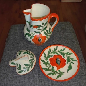 Może przedstawiać: Biały dzbanek ceramiczny z czerwono-zielonym wzorem kwiatowym. Dzbanek ma uchwyt i pokrywkę. Jest też pasujący talerz z tym samym wzorem.