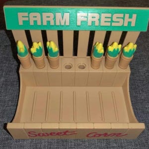 May include: A tan plastic toy farm stand with the words "FARM FRESH" in green and "Sweet Corn" in red. It features six toy corn cobs with yellow kernels and green husks.